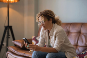 young woman seated on her couch at home and using her phone to answer the question, "Is medication-assisted treatment right for you?"