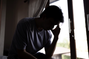 young man seated in dimly lit room next to a window with his head in his hands pondering the signs of meth addiction and how to recognize and seek help